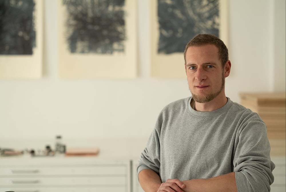 A professional portrait of a woman with shoulder-length dark hair and a warm smile, wearing a stylish blazer, standing in front of an art gallery.